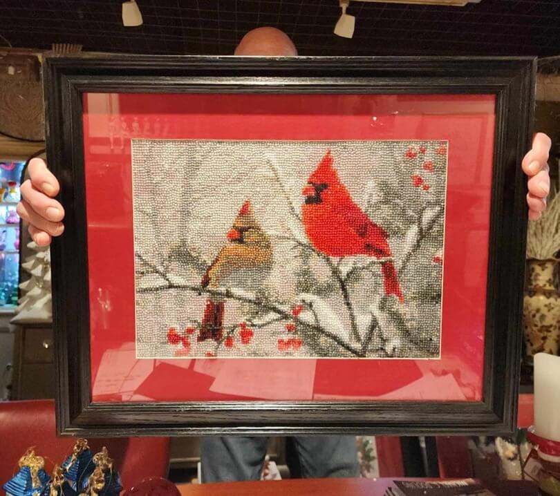 A man holding framed crystal diamond art in a black frame that shows a beautiful winter scene of a female and male cardinal looking into the distance